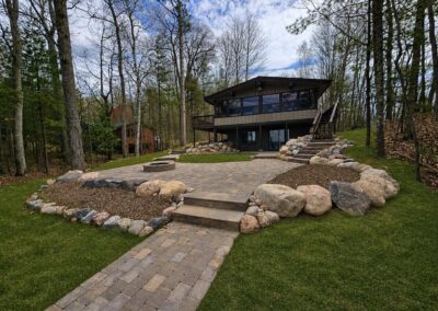Modern two-story house with large windows on a wooded lot, stone patio and fire pit.