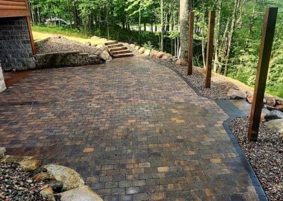 Curved brick-paver driveway with stone edging and wooden posts beside a forested yard.