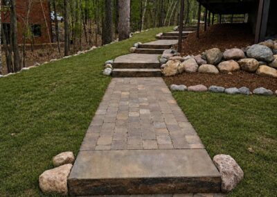 Stone brick pathway winding toward a raised wooden deck, with steps and a border of smooth rocks, flanked by green lawn and trees.”]} }{