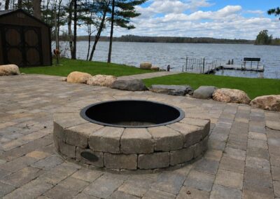 Circular stone fire pit built into a stone-paved patio overlooking a lake with a wooden dock in the distance and trees on the shore.