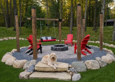Golden retriever resting on stone steps in front of a circular patio with a fire pit, red Adirondack chairs, and a wooden swing set in a forested yard.