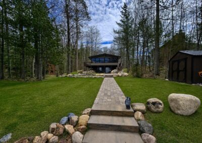 Stone walkway through a grassy yard leads to a two-story cabin with large windows in a forest setting.